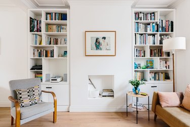 Modern living room with bookshelves, leather couch, chair, small side table, and small white fireplace underneath hung framed picture