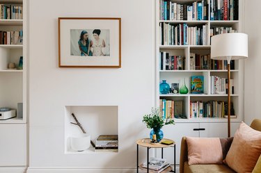 Modern living room with bookshelves, leather couch, lamp, small side table, and small white fireplace underneath hung framed picture