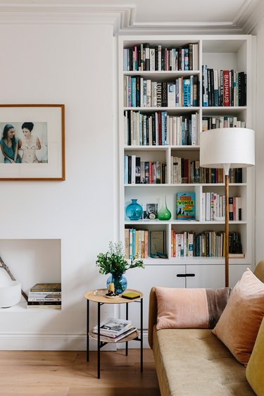 Modern living room with bookshelves, leather couch, lamp, small side table, and small white fireplace underneath hung framed picture