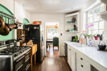 White cabinet kitchen with a white tile backsplash, black stove, white shelves, copper pans, flowers, and green accent wall.