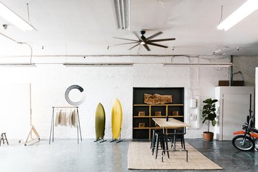 An industrial studio with surfboards, closet rack, wood table, a plant, ceiling fan, and fluorescent lights.
