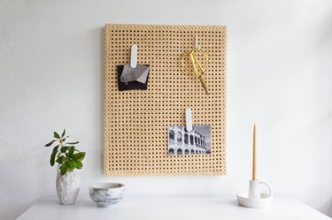 Cane memo board with scissors and photos over white desk with plant and candle