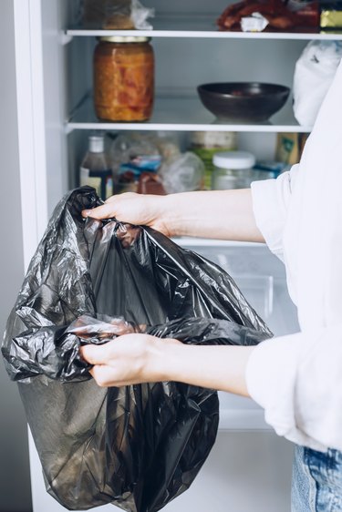 Hand holding black garbage bag in front of fully stocked white refrigerator