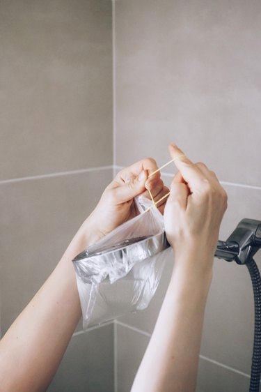 Hand attaching plastic bag to silver shower head against brown tiled shower