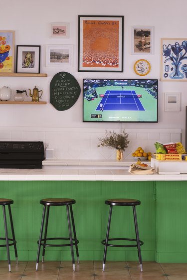 Kitchen bar setup with green tiled sides, white countertop, black stools, flat-screen TV, and sports parafernalia hung on pink wall