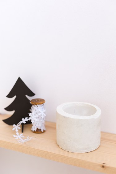 DIY concrete candle on wooden ledge next to snowflake string and Christmas tree toy against white background