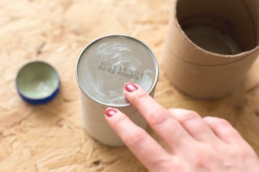 Hand checking wax in cylindrical container next to cap and other cylindrical container on wood tabletop