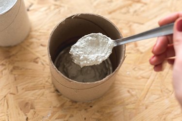 Hand mixing concrete with metal spoon in cylindrical cardboard container on wood countertop