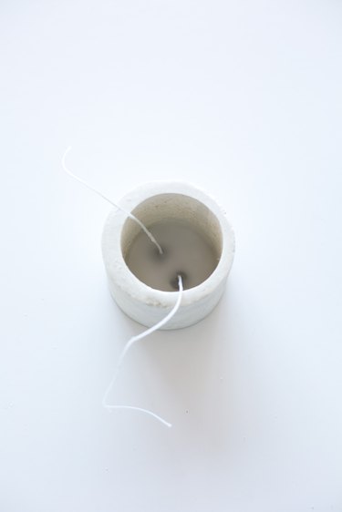 Two wicks partially submerged in wax in concrete candle holder on white background
