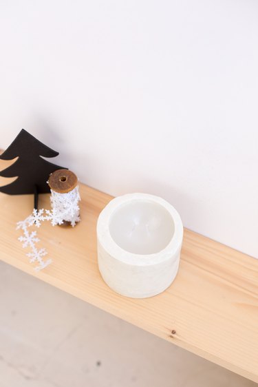 DIY concrete candle on wooden ledge next to snowflake string and Christmas tree toy against white background