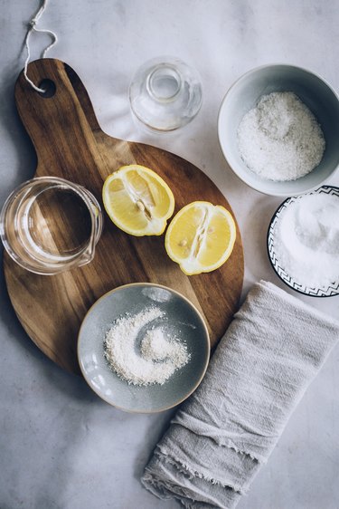 Lemons, round wood cutting board, and several small bowls with powdered soap resting on grey stone countertop