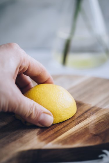 Hand rubbing lemon half on rounded wood cutting board against grey marble countertop