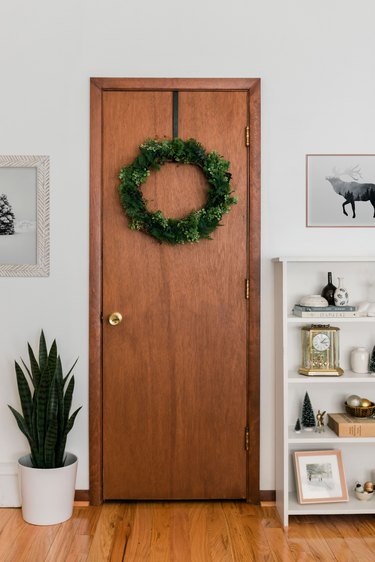 Wood door with faux evergreen Christmas wreath next to potted plant and bookshelf