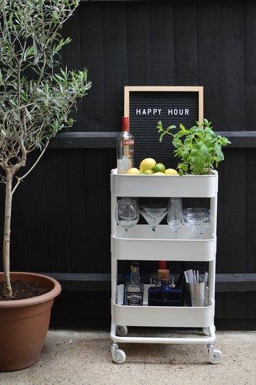 White bar cart with lemons, limes, plant, glasses, liquor bottles next to tree