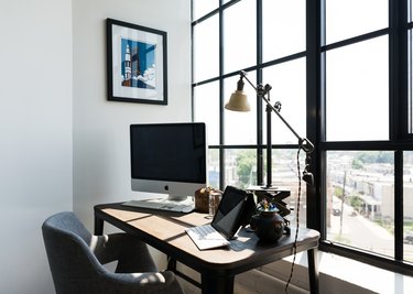 A wood desk with a computer and industrial lamp. A gray organic shaped chair. A large window with a black pane.
