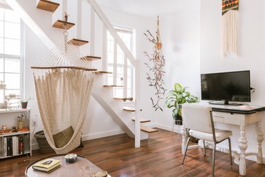 A white spindle leg desk with a desktop computer. Houseplant and textile art decor. A wood step staircase and wood flooring.