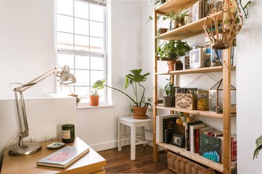 A wood bookcase with houseplants, books and art. A night table with a metal lamp and a book. A chair in the corner with a plant.