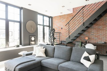 Contemporary upstairs space with grey couch, full-length windows, brick wall, and black metal staircase.