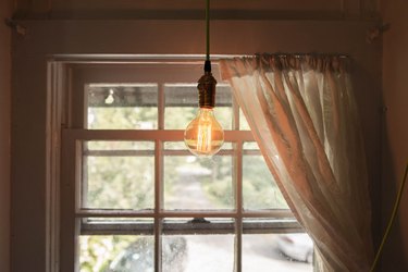 a bare edison bulb hangs in front of a window with a single gauzy curtain