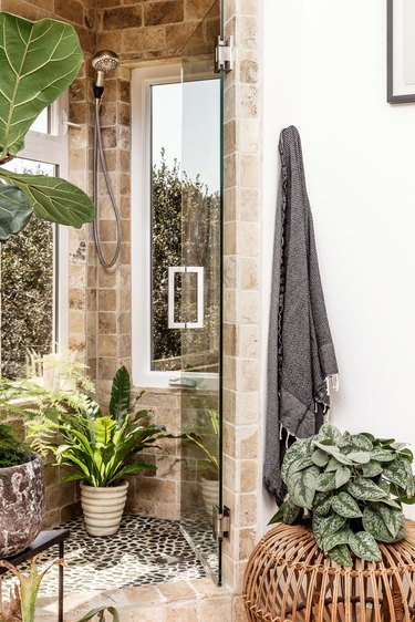 A shower with a mosaic pebble floor, rustic neutral tiles, windows, and glass shower door. Houseplants decorate the space.