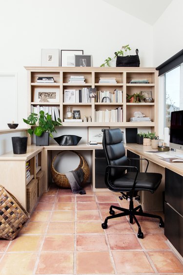 Office with a light wood desk and bookcase. Black chair, shades, cabinets and planters. Baskets and plants decorate the space.
