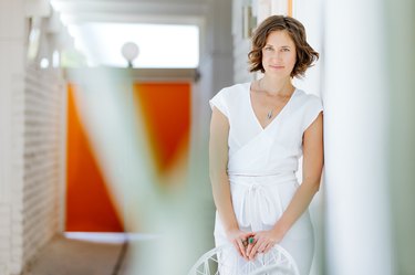 a woman in white leans on a white wall and holds a white chair