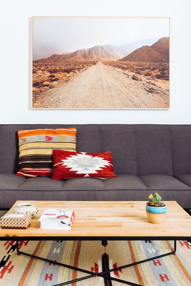 a desert landscape hangs above a a gray couch and a coffee table with a wooden top