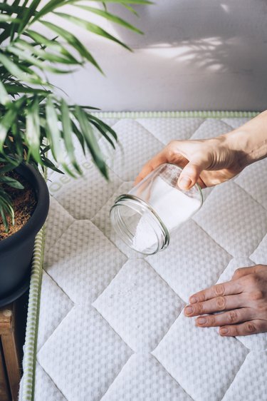 Hand pouring baking soda from small glass container onto white mattress next to medium potted plant in white room