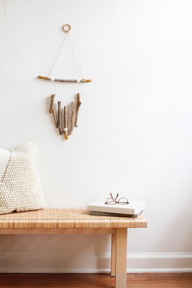 a wall hanging, made of several vertical pieces of driftwood suspended from a horizontal one, hangs above a wooden bench with a rattan top