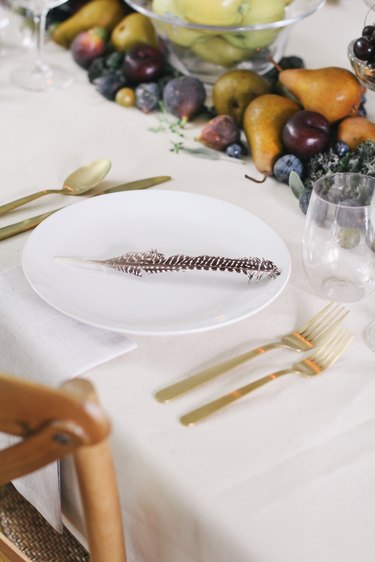 a table with a runner made of fruits and vegetables, gold cultery, and white plates with a single turkey feather on each one