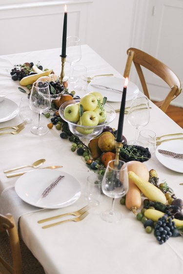 a line of produce, including grapes, pears, cherries, and squash runs down the length of a table with a white cloth and gold cutlery