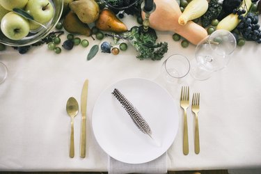 a table with a runner made of fruits and vegetables, gold cultery, and white plates with a single turkey feather on each one