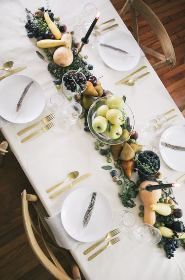 a line of produce, including grapes, pears, cherries, and squash runs down the length of a table with a white cloth and gold cutlery