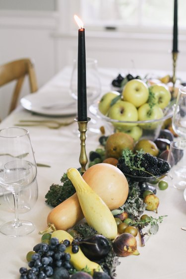 a line of produce, including grapes, pears, cherries, and squash runs down the length of a table with a white cloth and gold cutlery