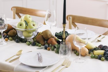 fall party idea with table runner of produce, including grapes, pears, cherries, and squash runs down the length of a table with a white cloth and gold cutlery