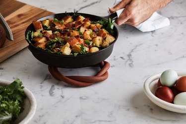 Cast iron pan with food on brown trivet with wood cutting board and dishes of food