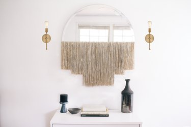 White fringe fringe mirror with gold sconces over dresser with vase