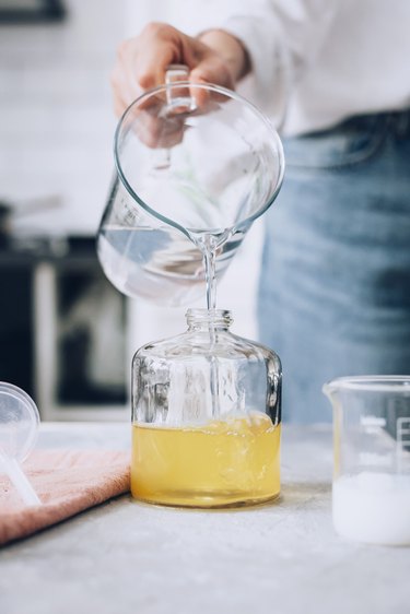 Hand pouring liquid from large measuring cup into mid-sized glass jar next to napkin and other glass containers on slate grey countertop