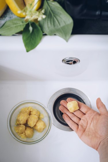 Hand holding lemon-filled garbage pod over garbage disposal with bowl filled with garbage pods in a white sink with black backsplash