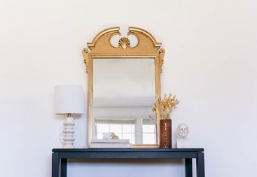 Gold antique style mirror on side table with lamp, vase of dried flowers and sculpture bust