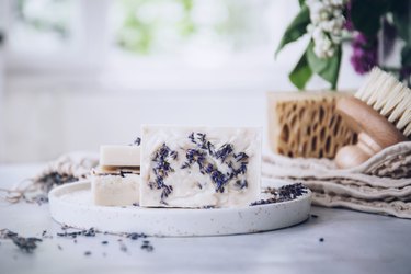 Goat milk soap bars covered with lavender in small white dish on white table next to round brush and tan sponge