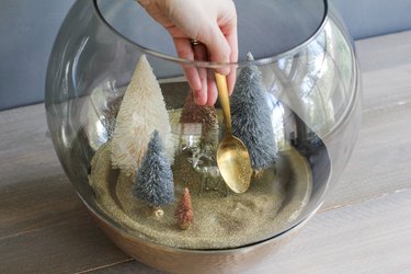 a crafter uses a gold spoon to add glitter to a holiday scene in a glass terrarium