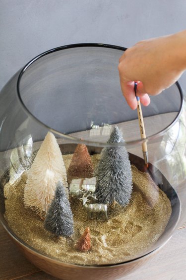 a crafter uses a paint brush to tidy up glitter inside a glass terrarium