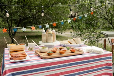 Picnic table with a red, white and blue tablecloth and star garland, hot dogs with condiments, and a condiment caddy.