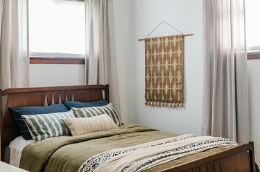 Brown patterned wall hanging rug next to bed with blue, pink, and green pillows against white background with windows on either side