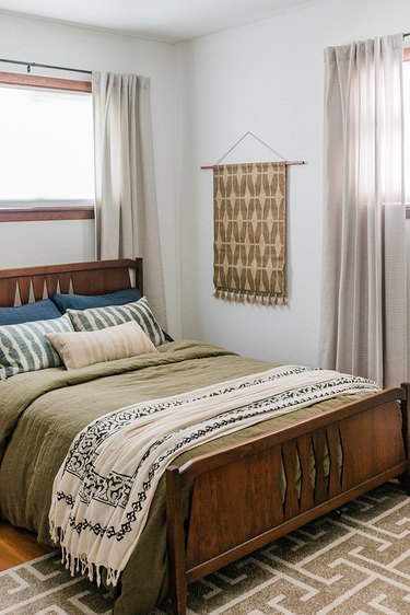 Brown patterned wall hanging rug next to bed with blue, pink, and green pillows against white background with windows on either side