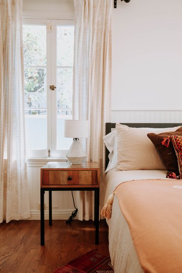 Casement window with curtains, wood nightstand with geometric lamp, a bed with brown-red pillows and white-peach bedding