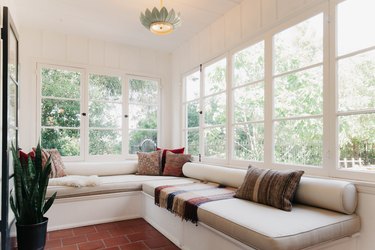 Sunroom with terra-cotta floor tile, white cushioned bench, multi-colored pillows, snake plant, and flower pendant light