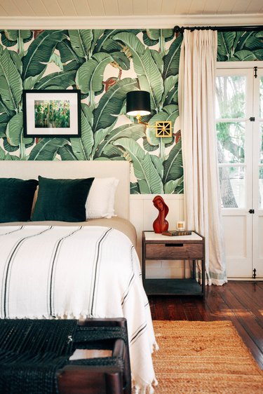 Bedroom with tropical palm wallpaper, white-green bedding, wood nightstand with sculpture, lamp sconce, and French door