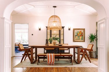 A dining room behind an archway. The walls are white and there is a long wood table surrounded by a bench and four wood chairs. There is a large woven pendant light on the ceiling and a floral painting on the wall.